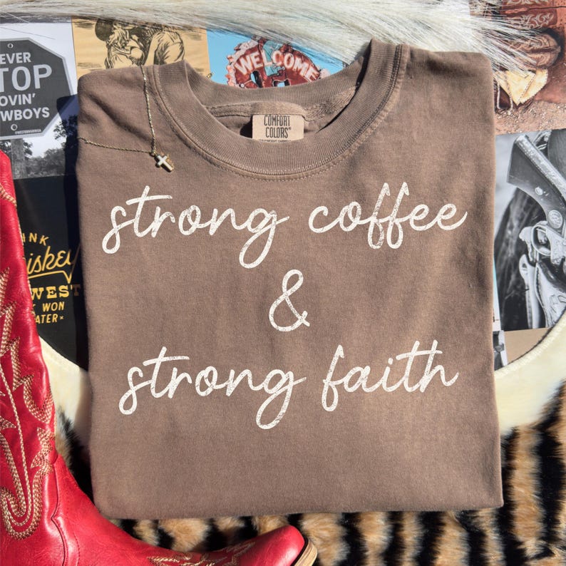 May include: A brown Comfort Colors sweatshirt with the words "strong coffee & strong faith" in white script. A gold cross necklace is visible. The sweatshirt is displayed on a patterned surface with a red boot.
