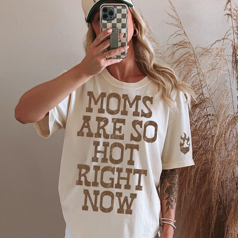 May include: Cream-colored t-shirt with the text "MOMS ARE SO HOT RIGHT NOW" in brown block letters. A small flame graphic is on the right sleeve. The shirt is a casual style.