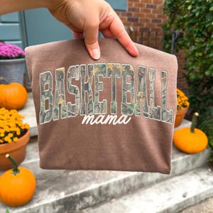 May include: A brown sweatshirt with the words "BASKETBALL mama" printed on the front. The text is in a camouflage pattern. The sweatshirt is being held up by a hand.