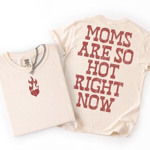 May include: Two cream-colored t-shirts. One shirt is folded, displaying a red flame graphic and a silver chain necklace. The other shirt has the text "MOMS ARE SO HOT RIGHT NOW" printed in red on the back. The shirts are on a white background.