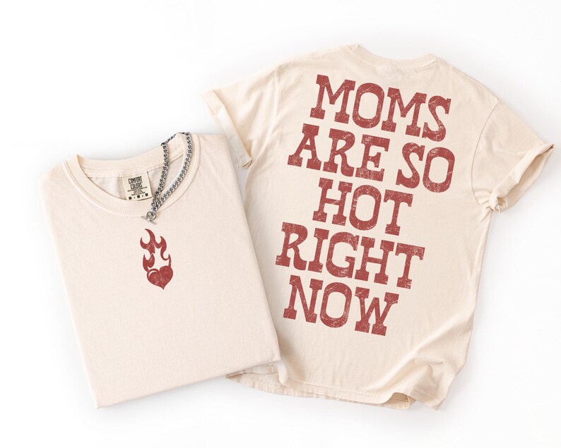 May include: Two cream-colored t-shirts. One shirt is folded, displaying a red flame graphic and a silver chain necklace. The other shirt has the text "MOMS ARE SO HOT RIGHT NOW" printed in red on the back. The shirts are on a white background.