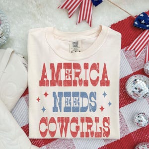 Może przedstawiać: Kremowa koszulka z napisem "AMERICA NEEDS COWGIRLS" w kolorze czerwonym i niebieskim. Koszulka otoczona jest patriotycznymi dekoracjami, w tym czerwonymi, białymi i niebieskimi wstążkami oraz kulami dyskotekowymi. Koszulka marki Comfort Colors.