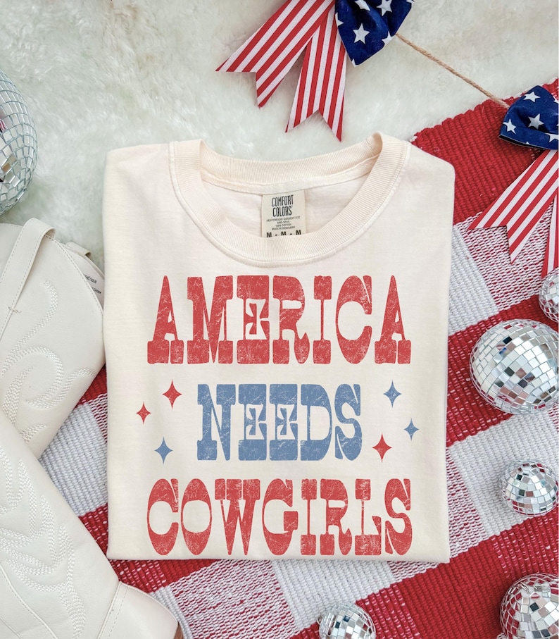 Może przedstawiać: Kremowa koszulka z napisem "AMERICA NEEDS COWGIRLS" w kolorze czerwonym i niebieskim. Koszulka otoczona jest patriotycznymi dekoracjami, w tym czerwonymi, białymi i niebieskimi wstążkami oraz kulami dyskotekowymi. Koszulka marki Comfort Colors.