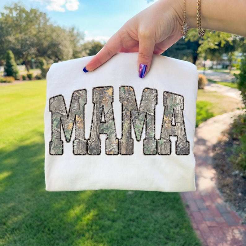 May include: White sweatshirt with the word "MAMA" in large, block letters. The letters have a camouflage pattern. The sweatshirt is held up against a background of green grass and trees.