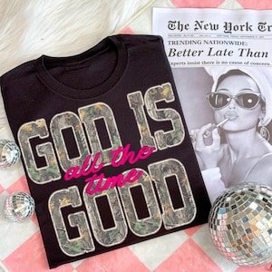 K&ouml;nnte beinhalten: Schwarzes T-Shirt mit dem Aufdruck "GOD IS GOOD all the time" in einem Tarn- und rosa Design. Das Shirt wird mit einer Zeitung und Discokugeln pr&auml;sentiert.