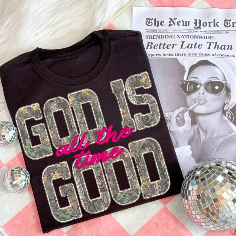 K&ouml;nnte beinhalten: Schwarzes T-Shirt mit dem Aufdruck "GOD IS GOOD all the time" in einem Tarn- und rosa Design. Das Shirt wird mit einer Zeitung und Discokugeln pr&auml;sentiert.