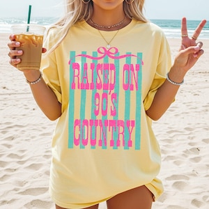 May include: A light yellow t-shirt with the text "RAISED ON 90'S COUNTRY" in pink and teal. The text is set against a striped background. The shirt is worn on a beach.