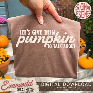 May include: A brown t-shirt featuring the white text LET'S GIVE THEM pumpkin TO TALK ABOUT. The shirt is held up against a background of pumpkins and flowers. The design is centered on the fabric, using a mix of bold and script fonts.