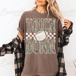 May include: A brown short-sleeve t-shirt featuring the text TOUCH DOWN in a camouflage pattern with a white football graphic and a checkered border. The shirt is worn with a black and tan plaid long-sleeve button-down shirt layered over it.