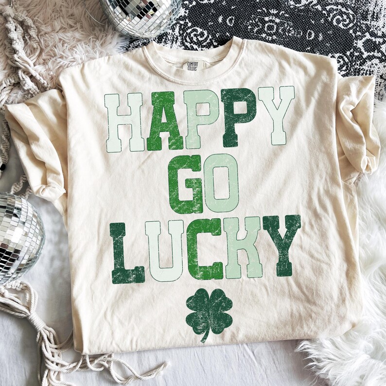 May include: Cream-colored t-shirt with the words "HAPPY GO LUCKY" in green lettering and a four-leaf clover graphic. The shirt has a relaxed fit and a crew neck.