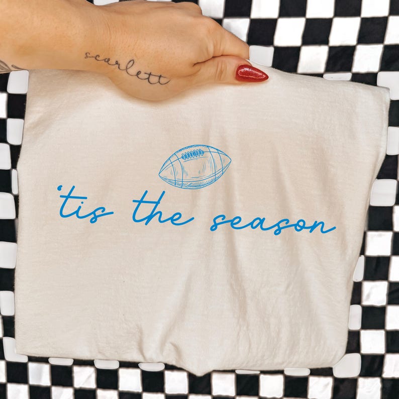 May include: A cream-colored t-shirt featuring a blue line-art illustration of an American football centered above the blue cursive text that reads 'tis the season. The design is printed on the front of the fabric, showcasing a simple graphic style.