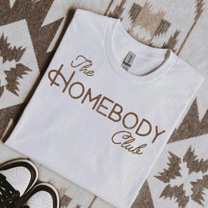 K&ouml;nnte beinhalten: Ein wei&szlig;es T-Shirt mit dem Text "The Homebody Club" in braunen Buchstaben. Das Shirt besteht aus einem weichen Material und liegt auf einer gemusterten Oberfl&auml;che. Ein Paar wei&szlig;er und brauner Turnschuhe ist unten links zu sehen.