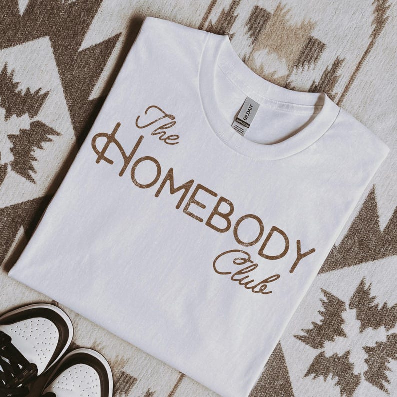 K&ouml;nnte beinhalten: Ein wei&szlig;es T-Shirt mit dem Text "The Homebody Club" in braunen Buchstaben. Das Shirt besteht aus einem weichen Material und liegt auf einer gemusterten Oberfl&auml;che. Ein Paar wei&szlig;er und brauner Turnschuhe ist unten links zu sehen.