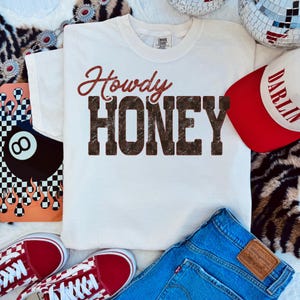 May include: A white t-shirt with the words "Howdy Honey" in a brown, distressed font. The image also includes a red and white baseball cap, blue jeans, and red sneakers. The overall theme is casual and country-inspired.