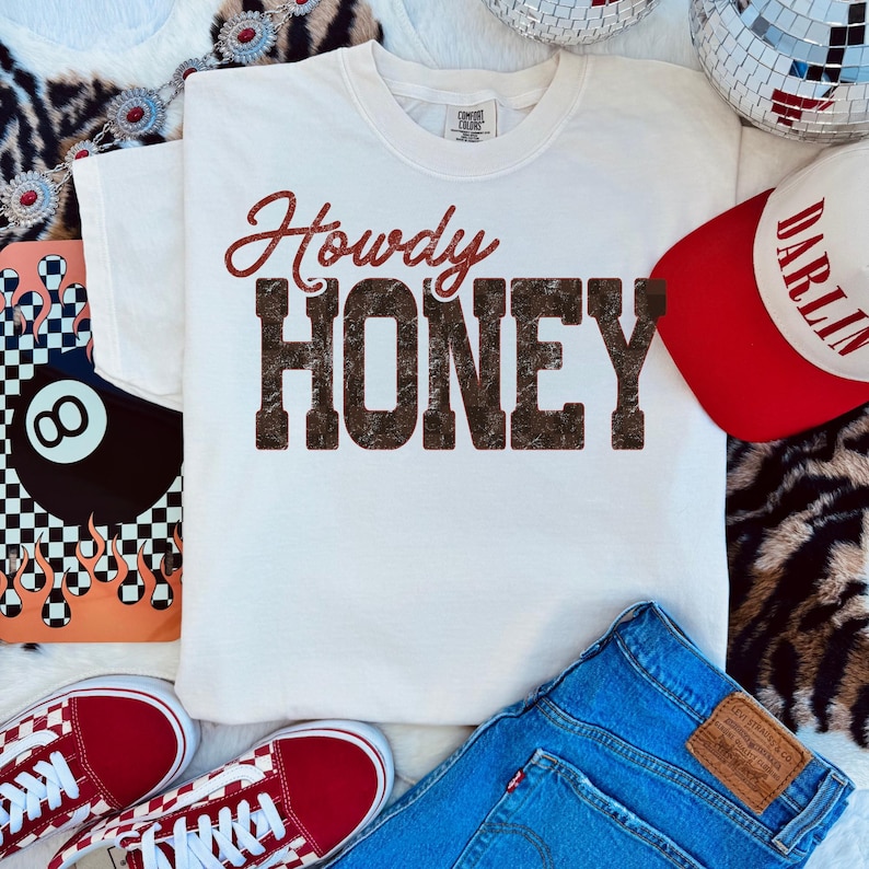 May include: A white t-shirt with the words "Howdy Honey" in a brown, distressed font. The image also includes a red and white baseball cap, blue jeans, and red sneakers. The overall theme is casual and country-inspired.