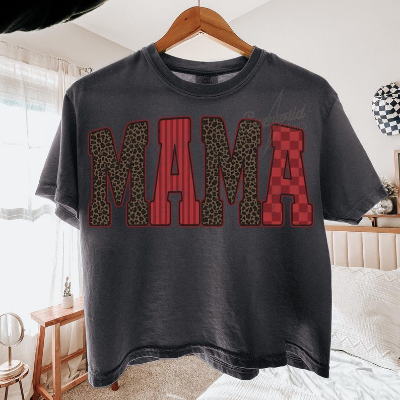 May include: A charcoal gray short-sleeve t-shirt featuring the word MAMA printed across the chest in large block letters. Each letter has a distinct pattern: the first and third letters have a brown leopard print, while the second and fourth letters feature red vertical stripes and a red checkered pattern respectively.
