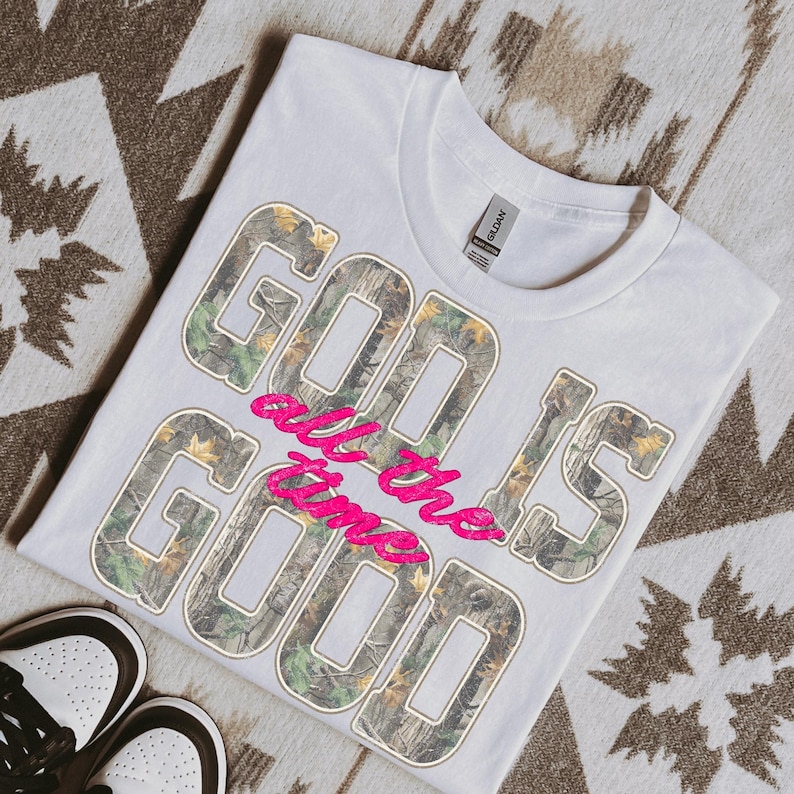 K&ouml;nnte beinhalten: Wei&szlig;es T-Shirt mit dem Text "GOD IS GOOD all the time" in einem Camouflage-Muster und pinkfarbener Schrift. Das Shirt liegt auf einer gemusterten Oberfl&auml;che, unten sind Turnschuhe zu sehen.