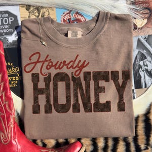 May include: A brown Comfort Colors sweatshirt with the words "Howdy Honey" in red and brown distressed lettering. A silver cross necklace rests on the shirt. The sweatshirt is surrounded by cowboy-themed items, including a red boot.