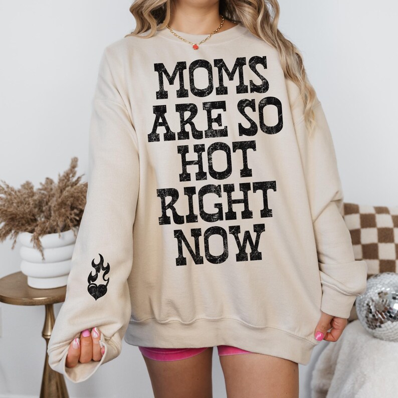 May include: A beige sweatshirt with the text "MOMS ARE SO HOT RIGHT NOW" in black block letters. A black heart with flames is printed on the left sleeve. The sweatshirt is a casual style.