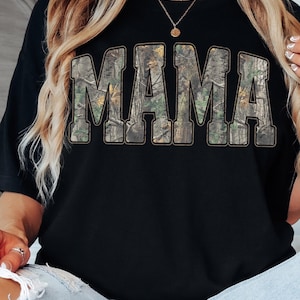 May include: Black t-shirt with the word "MAMA" in large, block letters. The letters are filled with a camouflage pattern. The shirt is worn by a person wearing a gold necklace and light blue jeans.