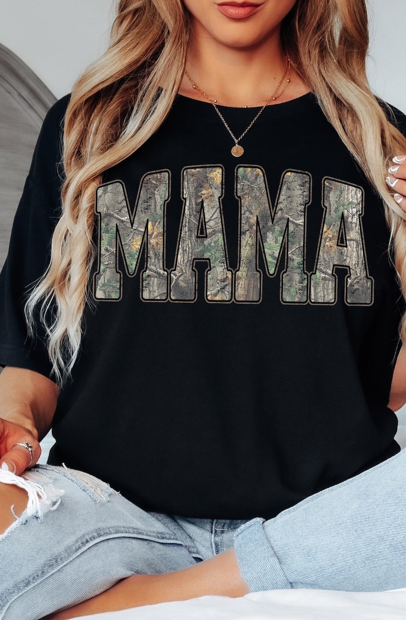 May include: Black t-shirt with the word "MAMA" in large, block letters. The letters are filled with a camouflage pattern. The shirt is worn by a person wearing a gold necklace and light blue jeans.