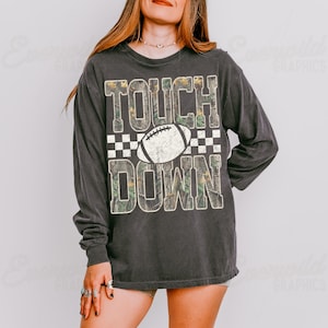 May include: A charcoal gray long-sleeve shirt featuring the text TOUCH DOWN in large block letters with a camouflage pattern. A white football graphic is centered between the words, and a black and white checkered pattern borders the text.
