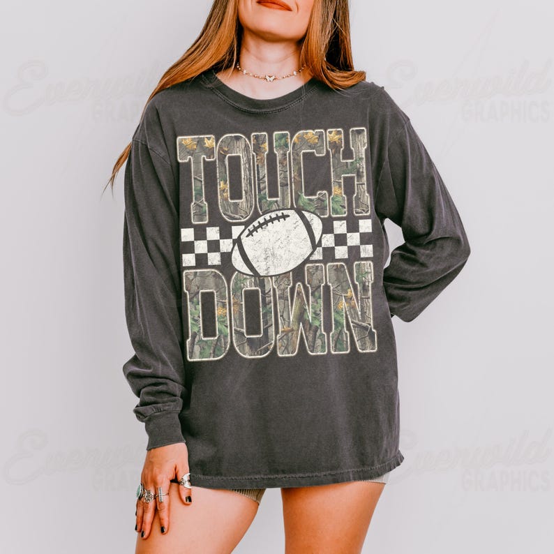 May include: A charcoal gray long-sleeve shirt featuring the text TOUCH DOWN in large block letters with a camouflage pattern. A white football graphic is centered between the words, and a black and white checkered pattern borders the text.