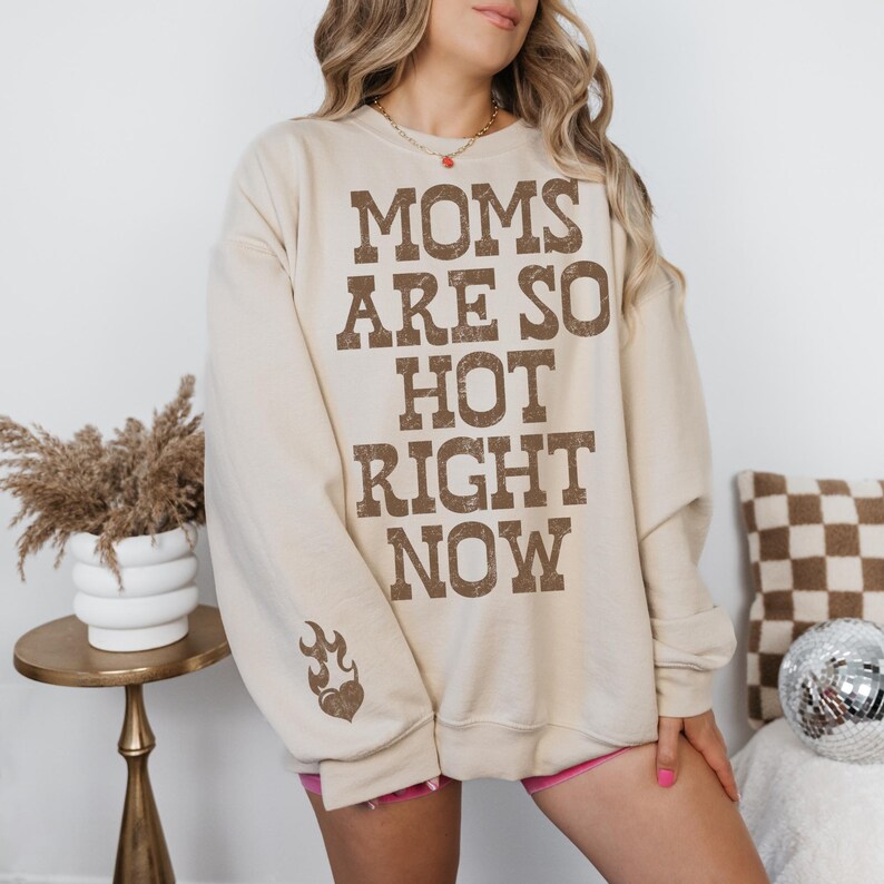 May include: A beige sweatshirt with the text "MOMS ARE SO HOT RIGHT NOW" in brown block letters. A flame and heart graphic is on the left sleeve. The sweatshirt is part of a clothing collection.
