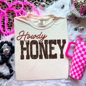 May include: Cream-colored t-shirt with the words "Howdy HONEY" in a distressed, brown font. The word "Howdy" is in a cursive, red font. The shirt is on a white, fluffy surface, with a pink checkered tumbler and disco balls.