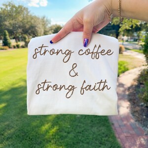 May include: A white t-shirt with the words "strong coffee & strong faith" printed in brown script. The shirt is held up against a background of green grass and a blue sky.