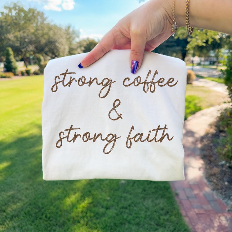 May include: A white t-shirt with the words "strong coffee & strong faith" printed in brown script. The shirt is held up against a background of green grass and a blue sky.