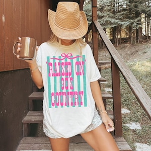 May include: Cream-colored t-shirt with the text "RAISED ON 90'S COUNTRY" in pink and teal stripes. The shirt is paired with a straw cowboy hat and a brown mug. The overall theme is country music and casual wear.