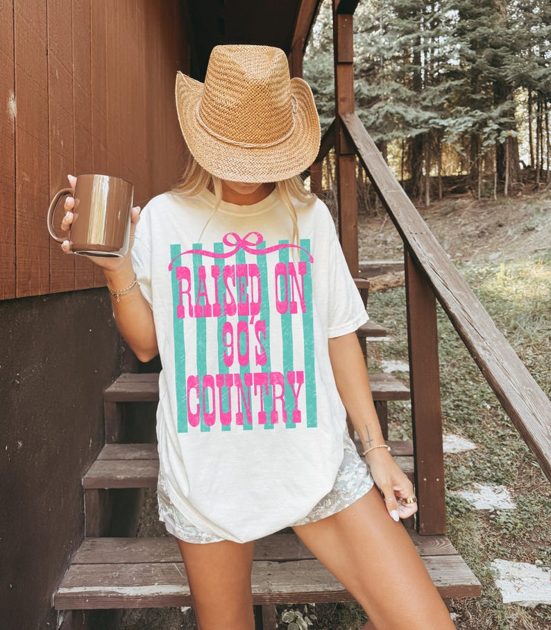 May include: Cream-colored t-shirt with the text "RAISED ON 90'S COUNTRY" in pink and teal stripes. The shirt is paired with a straw cowboy hat and a brown mug. The overall theme is country music and casual wear.