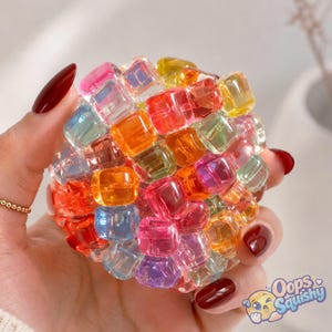 Peut inclure: Un jouet sensoriel sphérique composé de nombreux petits cubes en plastique translucide et multicolore dans des tons de rouge, orange, jaune, vert, bleu et rose. Les cubes sont disposés en une formation dense et groupée, créant une surface texturée.