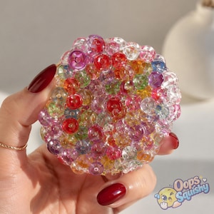 K&ouml;nnte beinhalten: Ein kreisf&ouml;rmiges, quetschbares sensorisches Spielzeug, das aus zahlreichen kleinen, durchscheinenden, bunten Kunststoffperlen in Rot, Gelb, Gr&uuml;n, Blau und Violett besteht. Die Perlen sind dicht gepackt und bilden eine texturierte, runde Oberfl&auml;che.
