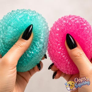 May include: Two round, textured scrubbers, one in translucent teal and one in bright pink, featuring a porous, bubbly surface structure. The scrubbers are held in place, showcasing their flexible, rounded shape and vibrant, semi-transparent colors.