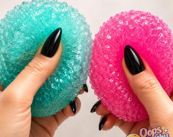 May include: Two round, textured scrubbers, one in translucent teal and one in bright pink, featuring a porous, bubbly surface structure. The scrubbers are held in place, showcasing their flexible, rounded shape and vibrant, semi-transparent colors.