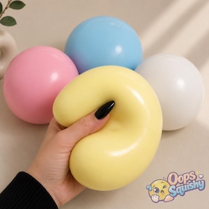 May include: A set of four smooth, round squishy toys in pastel colours including pink, light blue, white, and a yellow one being compressed. The yellow toy is being held and squeezed, showing its flexible texture. The text Oops Squishy is visible.