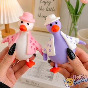 Puede incluir: Dos pequeñas figuras de pato hechas de material suave. Una es blanca con un sombrero rosa y una chaqueta rosa con estampado floral. La otra es morada con un sombrero blanco y una chaqueta blanca con estampado floral morado. Ambas tienen picos y patas naranjas.