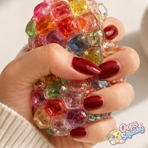 K&ouml;nnte beinhalten: Ein rundes, mehrfarbiges Squishy-Spielzeug, das aus zahlreichen kleinen, durchscheinenden, facettierten Kunststoffperlen in den Farben Rot, Gelb, Gr&uuml;n, Blau und Violett besteht. Das Spielzeug wird gedr&uuml;ckt, der Text "Oops Squishy" ist sichtbar.