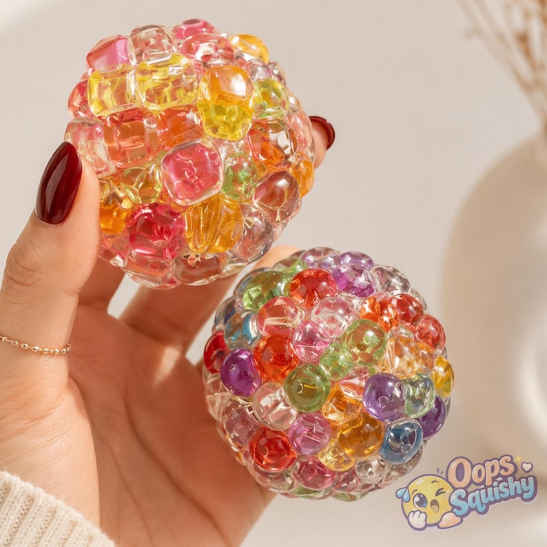 K&ouml;nnte beinhalten: Zwei kugelf&ouml;rmige Stressb&auml;lle aus transparentem, flexiblem Material, gef&uuml;llt mit kleinen, mehrfarbigen, durchscheinenden Perlen in Rot, Gelb, Orange, Gr&uuml;n, Violett und Blau. Der Text "Oops Squishy" erscheint in der unteren rechten Ecke.