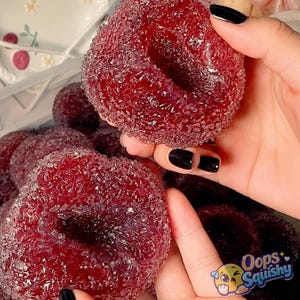 Può includere: Una collezione di giocattoli morbidi, rotondi e di colore rosso intenso, con una superficie strutturata, porosa e cristallina. Ogni oggetto presenta un'indentazione circolare centrale, che ricorda la forma di una ciambella. Il materiale appare soffice.
