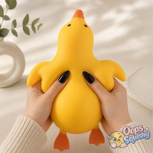 May include: A bright yellow duck-shaped squishy toy with an orange beak and feet, small black eyes, and a smooth texture. The toy is being squeezed, showing its flexible material. The text "Oops Squishy" with a small cartoon face is visible.