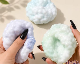 May include: Three round, textured squishy toys with a bumpy, cloud-like surface. One is light blue, one is a pale lavender, and one is a soft mint green. The text "Oops Squishy" with a small cartoon face is visible in the bottom right corner.