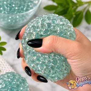 May include: A round, translucent, light green squishy ball filled with numerous small, clear, bead-like spheres. The texture is bumpy and uneven due to the internal beads. The text "Oops Squishy" is printed in the bottom right corner of the image.