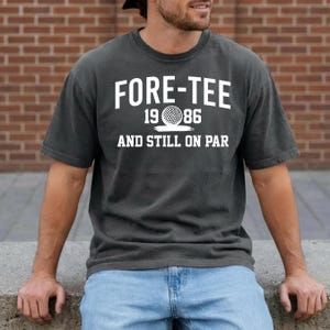 Może przedstawiać: Ciemnoszara koszulka z białym napisem "FORE-TEE 1986 AND STILL ON PAR". Projekt zawiera grafikę piłki golfowej. Koszulka jest w stylu casual.