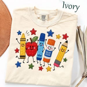 Op de afbeelding: Een ivoorkleurig t-shirt met een geborduurde afbeelding van een gele waskrijt, een rode appel, een blauwe stift, een oranje lijmstift met het label "GLUE STICK" en een geel potlood, allemaal met lachende gezichten en omringd door kleine gekleurde sterren.