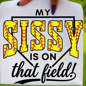 May include: White t-shirt with the text "MY SISSY IS ON that field!" in black and yellow. The word "SISSY" is designed to resemble a baseball with red stitching. The shirt is held up against a green background.