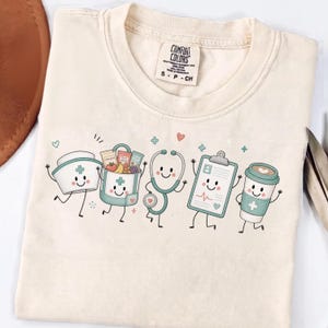 Op de afbeelding: Een crèmekleurig t-shirt met een Comfort Colors merklabel in de nek. De voorkant toont een afbeelding van vijf lachende, antropomorfe medische voorwerpen: een verpleegsterskapje, een lunchtas, een stethoscoop, een klembord en een koffiebeker.