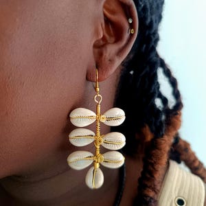 Puede incluir: Pendientes de alambre dorado con conchas blancas colgando de ellos. Los pendientes son colgantes y tienen un estilo bohemio.